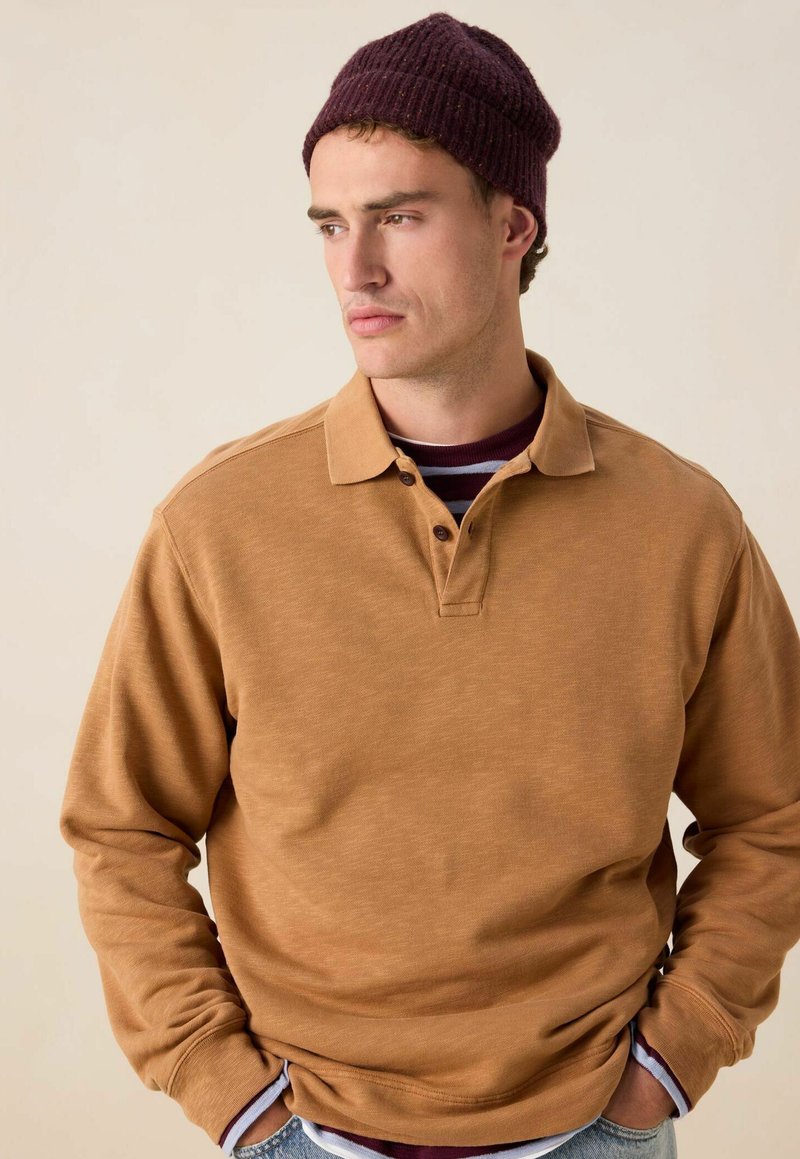 Bruine lange mouwen polo met een kraag, voorzien van een sluiting met drie knopen. Daaronder steekt een bordeaux gestreept shirt naar voren. Donkere gebreide muts op het hoofd.