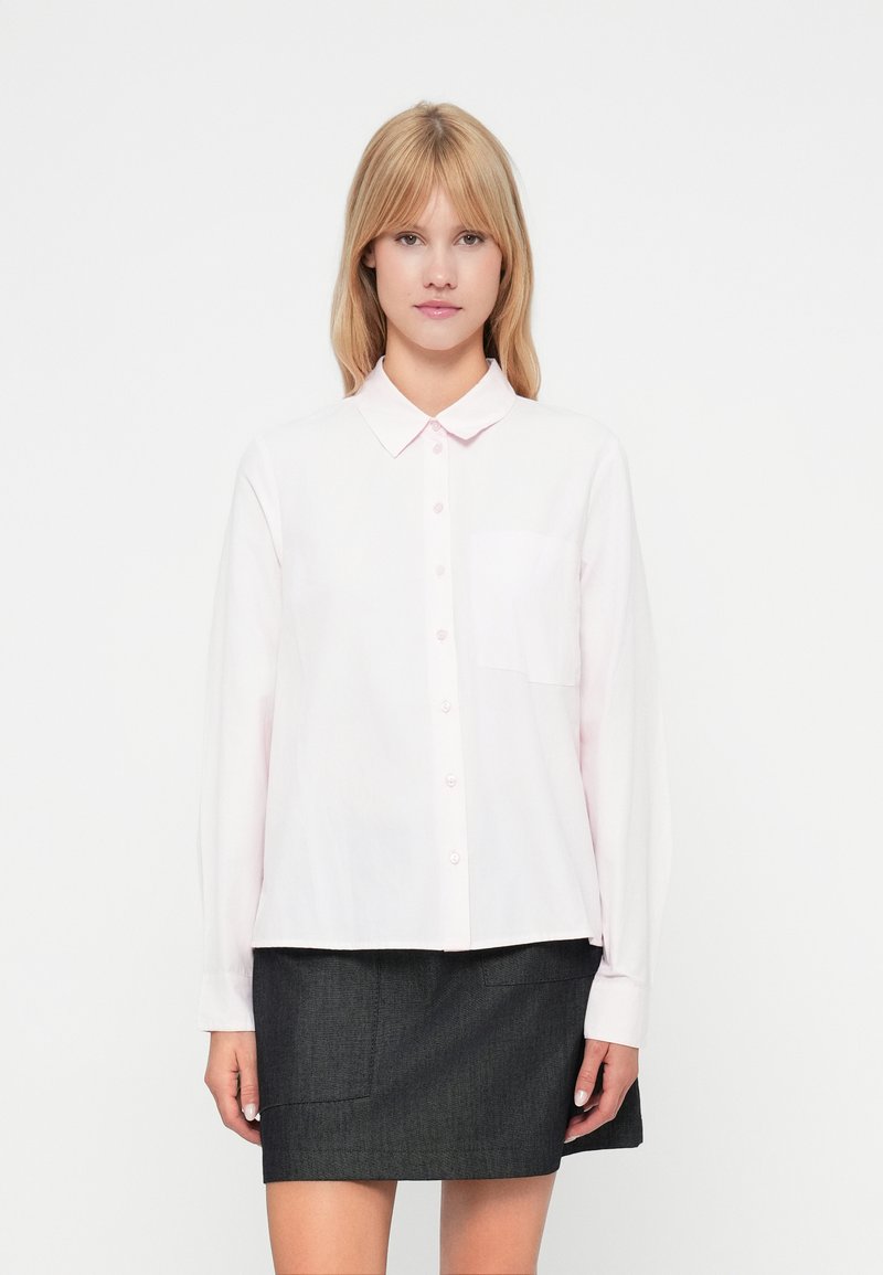 Mulher loira vestindo uma camisa de botão de mangas longas em rosa claro com um bolso no peito e uma saia mini cinza escura, de pé contra um fundo branco.