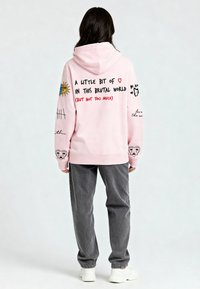 Ružičasta dukserica s kapuljačom s grafičkim tekstom i crtežima na leđima, prikazujući sunce, srca i apstraktne dizajne. Kombinirana s sivim hlačama i bijelim tenisicama.