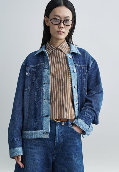 Jeansjacke mit hellblauem Kragen und dunkelblauem Körper, mit Knopfverschluss, getragen über einem braun-weiß gestreiften Hemd, kombiniert mit dunkelblauen Hosen.