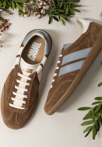 Baskets en daim marron avec des rayures bleu clair et des lacets blancs, entourées de feuilles vertes et de petites fleurs blanches.