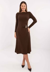 Brązowa sukienka z długim rękawem, dopasowaną górą, płynną spódnicą oraz złotym akcentem na lewym ramieniu i talii. Gładka tekstura.