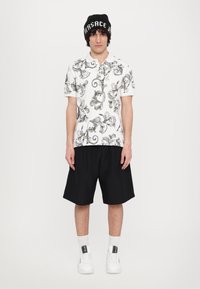 Biely polokošeľa s čiernym kvetinovým vzorom, čierne šortky a biele tenisky. Čierna čiapka s bielym textom. Jednoduchý, neformálny outfit.