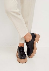 Chaussures en cuir noir avec des semelles en caoutchouc beige épaisses, présentant un bout rond, des détails de lacets et un design de semelle rugueux et texturé.