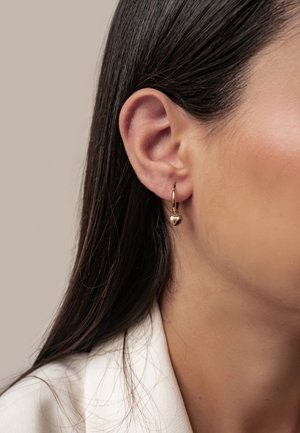 Nahaufnahme vom rechten Ohr einer Frau mit glatten dunklen Haaren, die einen kleinen goldenen Creolen-Ohrring mit einem Herzanhänger trägt.