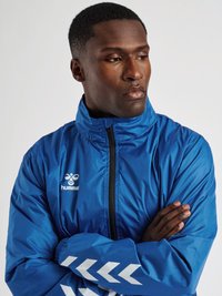 Mand i blå Hummel sportjakke med hvid chevron og logo, der ser til siden med armene over kors mod en ensfarvet baggrund.