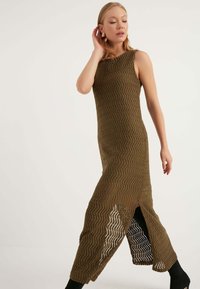 Abito lungo in maglia verde oliva con un motivo geometrico a zigzag, lati aperti e una silhouette aderente. Senza maniche, lungo fino al pavimento con uno spacco laterale.