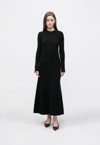 Czarna sukienka z długim rękawem wykonana z gładkiej, elastycznej tkaniny, z dopasowanym gorsetem i rozkloszowanym dołem. Noszona z bordowymi butami na obcasie.