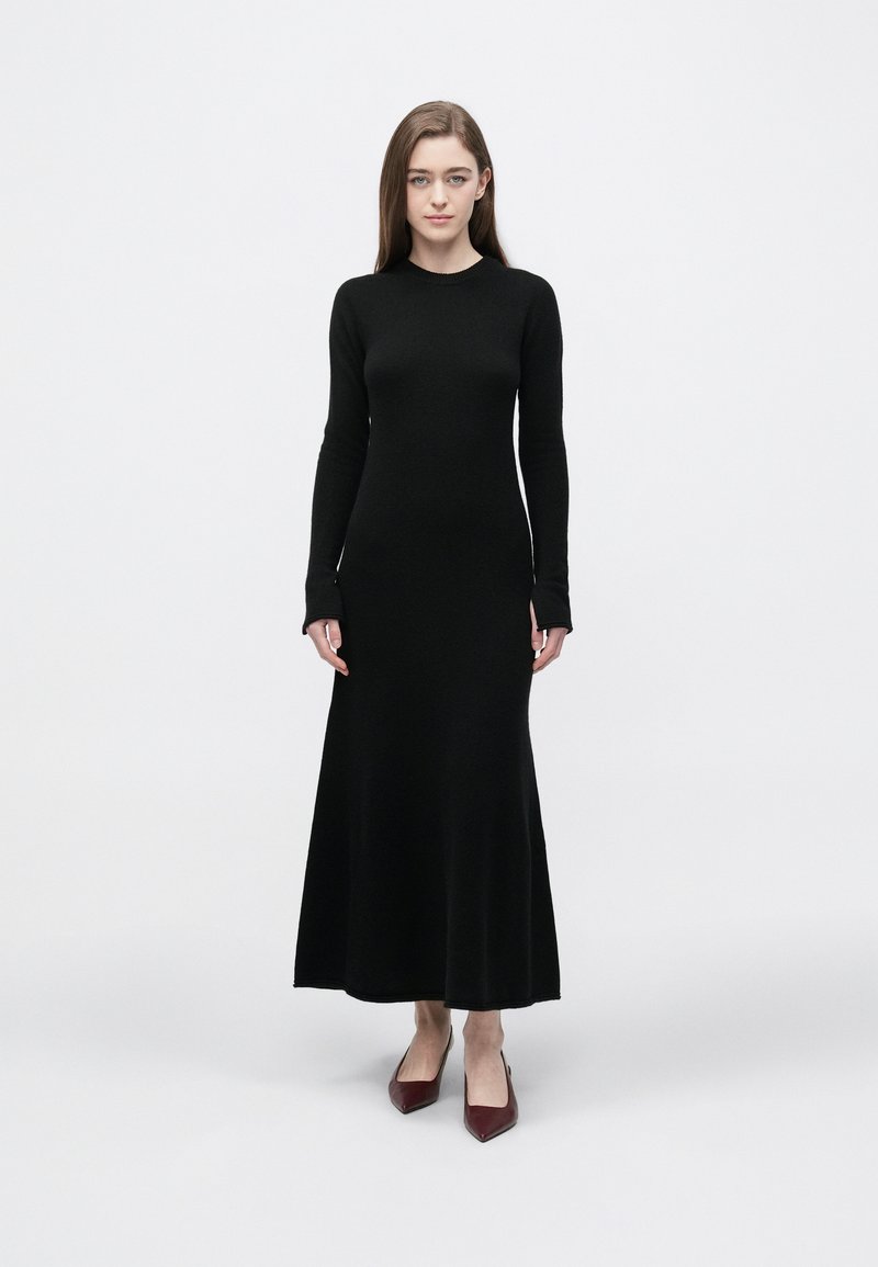 Czarna sukienka z długim rękawem wykonana z gładkiej, elastycznej tkaniny, z dopasowanym gorsetem i rozkloszowanym dołem. Noszona z bordowymi butami na obcasie.