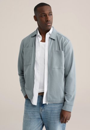 Lichtblauwe jas met ritssluiting en geribbelde textuur, twee borstzakken en een kraag, gedragen over een wit shirt en licht denim jeans.