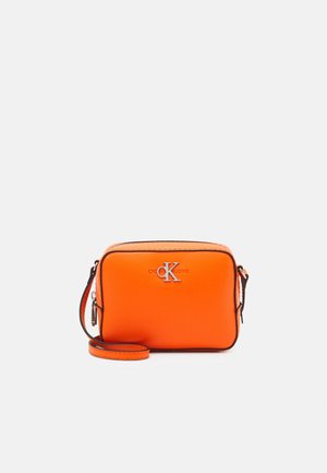 Kompaktowa, jasnopomarańczowa torba na ramię z czarnym wykończeniem i srebrnym logo „CK” na froncie, wyposażona w zamek błyskawiczny i regulowany pasek.