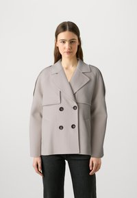 Cappotto grigio doppiopetto realizzato in tessuto morbido con ampi rever, due tasche frontali e bottoni neri. Indossato sopra jeans scuri.