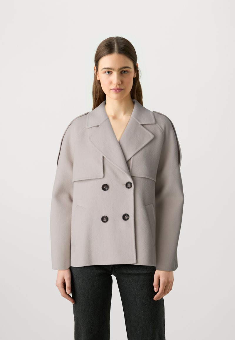 Cappotto grigio doppiopetto realizzato in tessuto morbido con ampi rever, due tasche frontali e bottoni neri. Indossato sopra jeans scuri.