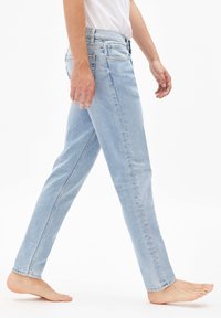 Jasnoniebieskie dżinsy z denimu o prostym kroju, z pięcioma kieszeniami i subtelnym blaknięciem. Model chodzi boso, prezentując dopasowanie spodni.