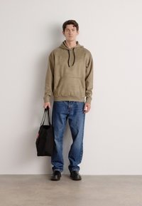 Hoodie verde-oliva com um bolso canguru, combinado com jeans azuis e sapatos pretos. A pessoa segura uma bolsa de lona preta.