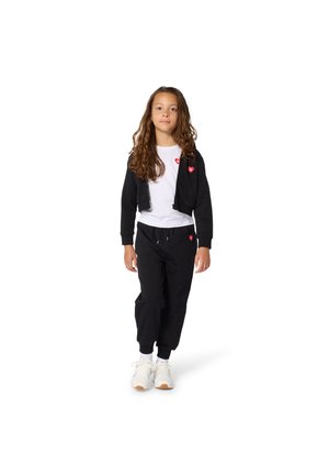 Zwarte hoodie met ritssluiting en rood hartlogo, gedragen over een wit T-shirt en zwarte joggingbroek. Het model draagt witte sneakers met een beige zool.