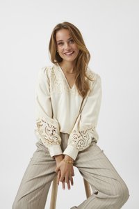 Blusa di crema con dettagli in pizzo intricati su spalle e maniche, abbinata a pantaloni a righe grigio chiaro. Sgabello di legno sullo sfondo.