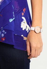 Okrągły zegarek na rękę z jasną, drewnianą tarczą, białym teksturowanym paskiem i metalowymi akcentami. Zestawiony z fioletową kwiatową bluzką i ciemnymi dżinsami.