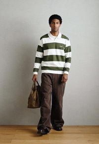 Rohelise ja valge triibulise pikade varrukatega polosärk, pruunid laiade säärtega püksid, kandes pruuni kotti; madalad kingad, seisab neutraalse tausta ees.