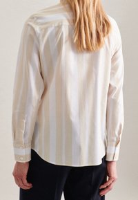 Person with long blonde hair wearing a beige and white vertical striped long-sleeve shirt and dark pants, shown from the back.