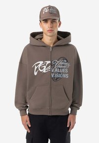 Brauner Kapuzenpullover mit Reißverschluss und Fronttaschen, versehen mit weißem und blauem grafischem Text. Kombiniert mit passender Mütze. Weiche Textur und lockere Passform.