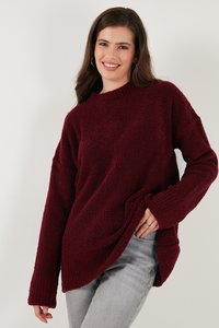 Donna sorridente con lunghi capelli castani indossa un maglione rosso scuro testurizzato e jeans grigi chiari, in posa su uno sfondo chiaro e uniforme.