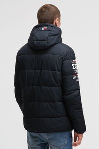 Man draagt een zwarte gewatteerde jas met capuchon en witte letters op de rechtermouw, kijkt weg tegen een effen achtergrond.