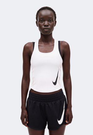 Vrouw die een witte mouwloze Nike top en zwarte Nike shorts draagt, staand tegen een effen lichte achtergrond.