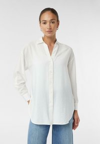 White button-up shirt with a collar, long sleeves, and a relaxed fit. Features a textured fabric and side pockets. Paired with blue jeans.