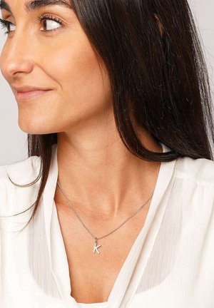 Silberne Kettenhalskette mit einem kleinen weißen Anhänger in Form des Buchstabens "K". Modell trägt eine hellfarbige, strukturierte Bluse.