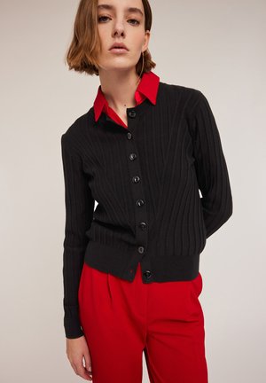 Schwarzer gerippter Strickcardigan mit Knopfverschluss, darunter ein rohes Hemd mit Kragen. Getragen mit roten Hosen aus strukturiertem Stoff.