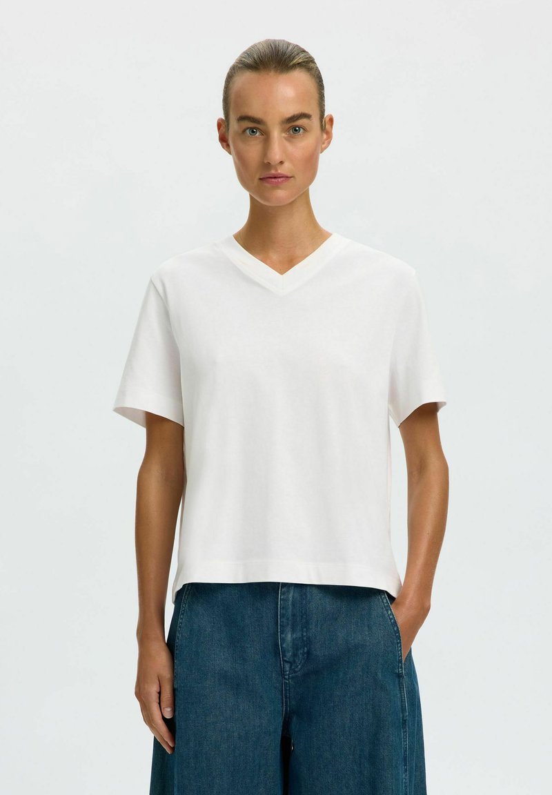 Femme avec les cheveux attachés en arrière portant un t-shirt blanc uni à manches courtes et col en V et un jean bleu, une main dans la poche sur un fond clair.