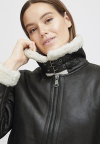 Jachetă din piele neagră cu un guler căptușit din blană albă, închidere cu fermoar și două curele cu catarame argintii la gât. Textură netedă.