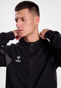 Felpa nera con zip e dettagli grigi, presenta un logo sul petto, polsini a coste e una superficie di tessuto liscia.