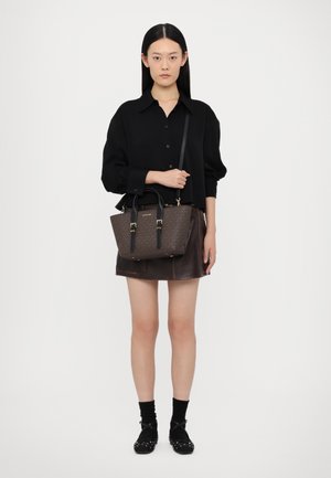 Braune Handtasche mit Muster und schwarzen Ledergriffen, goldenen Hardware-Akzenten. Das Modell trägt ein schwarzes Shirt und einen Rock aus braunem Leder.