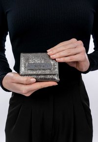 Cartera plateada metálica con acabado texturizado y forma rectangular. Cuenta con un detalle negro liso con el nombre de la marca.