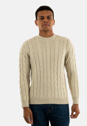 Hombre con un suéter de punto beige y jeans azul oscuro, de pie contra un fondo blanco liso.