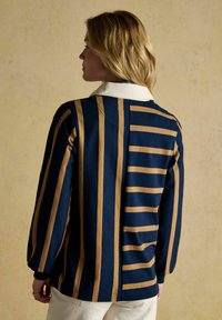 Woman facing away wearing navy blue jacket with vertical and horizontal tan stripes and white collar, beige pants, standing against plain background.