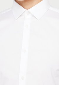 Lähikuva valkoisen nappikauluspaidan kauluksesta ja ylänapeista yksinkertaisella valkoisella taustalla.