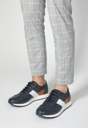Navy leather sneakers with grey mesh accents, white stripe detail, and brown leather patch. Rounded toe and rubber sole. Worn with checkered trousers.
