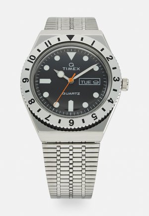 Quartz wristwatch with a black dial, silver case, and metal mesh band. Features white hour markers, orange second hand, and day-date display.