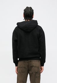 Felpa nera con cappuccio in tessuto strutturato con vestibilità comoda, abbinata a pantaloni marroni con più tasche e dettagli di cuciture.