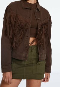 Bruine spijkerjack met lange franjes op de schouders, knoopsluiting aan de voorkant en een kort model, gecombineerd met een groene minirok.