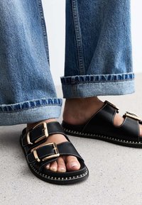 Sandali in pelle nera con due fasce larghe e fibbie dorate. Dettaglio di cucitura in avorio lungo la suola. Abbinati a jeans blu risvoltati.