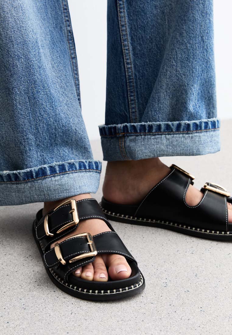 Sandali in pelle nera con due fasce larghe e fibbie dorate. Dettaglio di cucitura in avorio lungo la suola. Abbinati a jeans blu risvoltati.