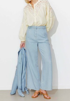 Femme portant un chemisier crème à panneaux en dentelle avec manches bouffantes, jean large taille haute bleu clair, sandales marron, et tenant une veste en jean.