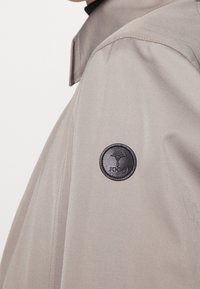 Cappotto beige in tessuto liscio e testurizzato, con una patch circolare nera sulla spalla e un colletto standard.