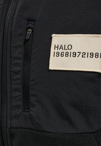 Svart zip-up-jacka med strukturerad tyg, sidofack med dragkedja och en rektangulär vävd lapp med "HALO" och siffror i svart tryck.