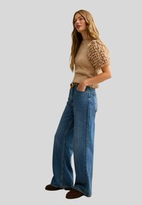 Knit sweater in beige with textured puff sleeves, paired with high-waisted, wide-leg blue denim jeans. Model stands against a grey background.