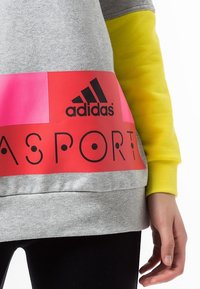 Sweat-shirt gris avec accents roses et rouges, arborant le logo Adidas et le texte "SPORT". Manches longues jaunes et ourlet côtelé.